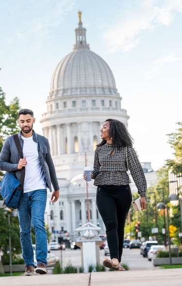 A man and woman walk away from the Wisconsin State Capitol toward the Monona Terrace. The main has a laptop bag over his shoulder and the woman is carrying a cup of coffee.