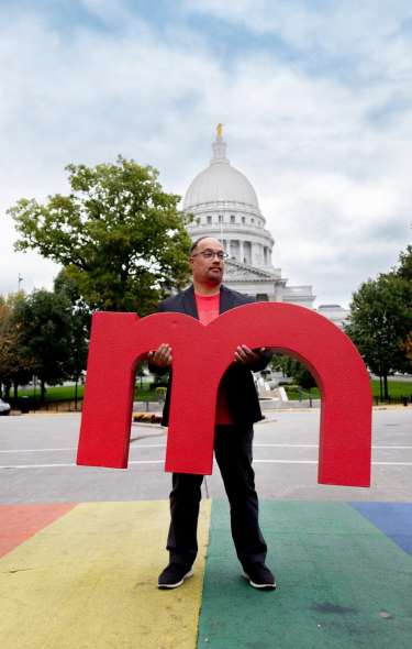 A man holds a large red M in front of the Wisconsin State Capitol while standing on a painted rainbow road