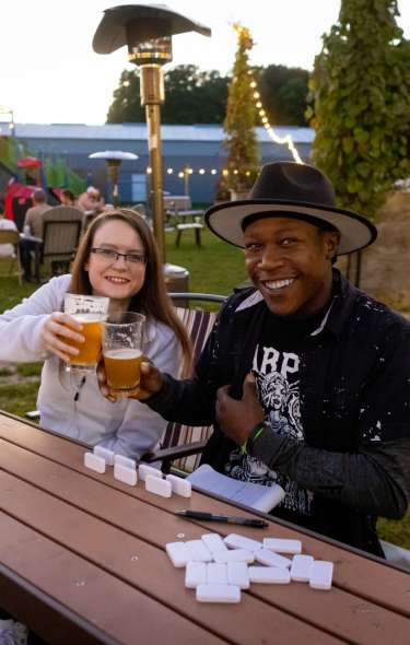 A Black man and a white woman cheers beer glasses while playing dominoes on a picnic bench at the Doundrins Distilling biergarten