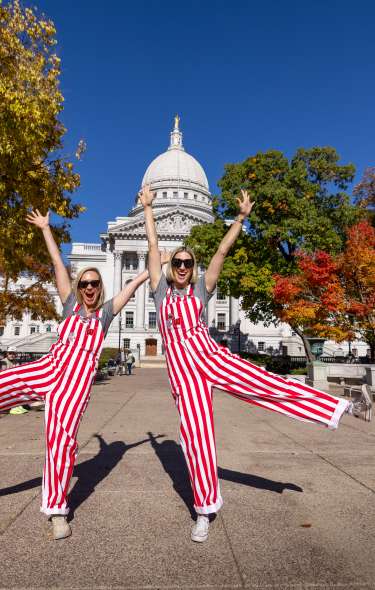 Two women wearing red and white striped overalls posing in front of the Wisconsin State Capitol building.
