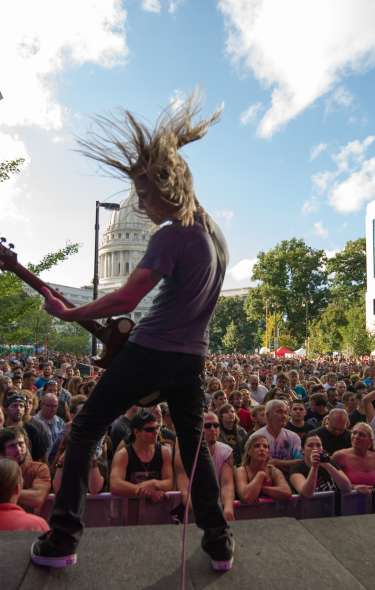 A wide view of a crowd of people watching a concert downtown from the perspective of the stage. A guitarist with long hair plays for the crowd