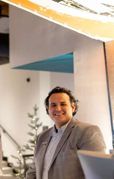A man smiles to the camera from behind the check in desk at Hotel Indigo. His coworker next to him is talking on the phone.