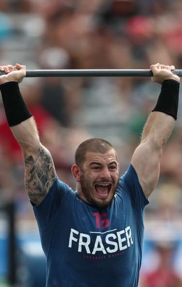 A man lifts a barbell with heavy weights on it above his head. He is yelling and there is a crowd of people watching in the background