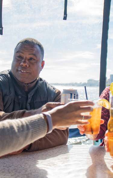 A group of four older people, one Black woman, one Black man, one white woman and one white man, clink cocktail glasses and beer bottles on a boat.