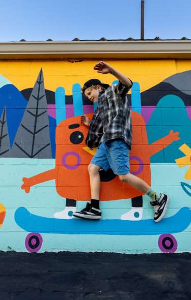 A young white boy jumps in front of a colorful mural, which makes it look like he is riding a skateboard