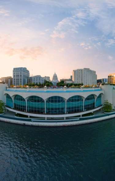 A panoramic view of a waterfront cityscape at sunset, featuring modern buildings and a serene lake. The sky is vibrant with colors.
