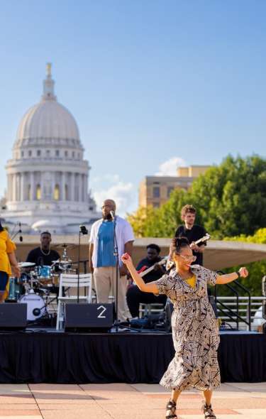A band plays on the Monona Terrace rooftop as a woman dances, including the Capitol and Monona Terrace at night