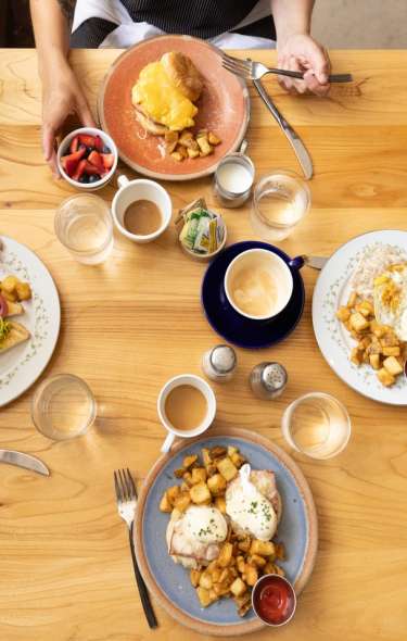 A wooden table displays a variety of breakfast dishes, including eggs, potatoes, and fruit. Coffee cups and condiments are also present.