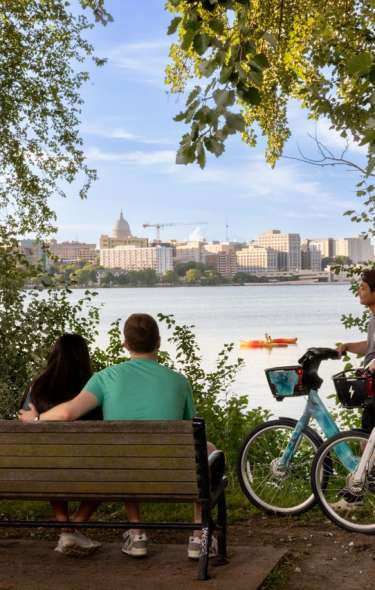 Four people sit on a bench next to two people on Bcycle e-bikes overlooking Lake Monona with the isthmus and Capitol Building in the background.