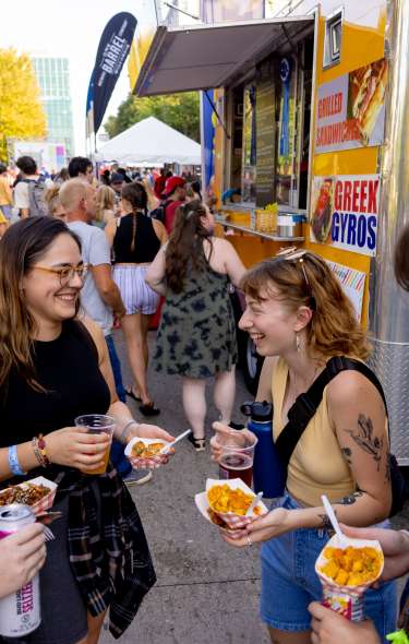 People gather beside a yellow food truck at an outdoor food festival, holding food and drinks while a crowd lines up along the tree-lined street.