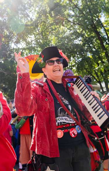 Three people wearing red coats and funky hats, holding various instruments as they walk and perform in a parade.