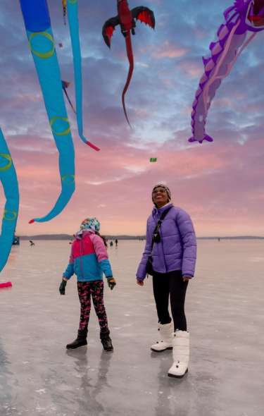 Two people, a Black woman and a child, stand on a frozen lake, gazing up at colorful kites soaring in the sky. The scene features vibrant designs, including dragons and long, flowing shapes.