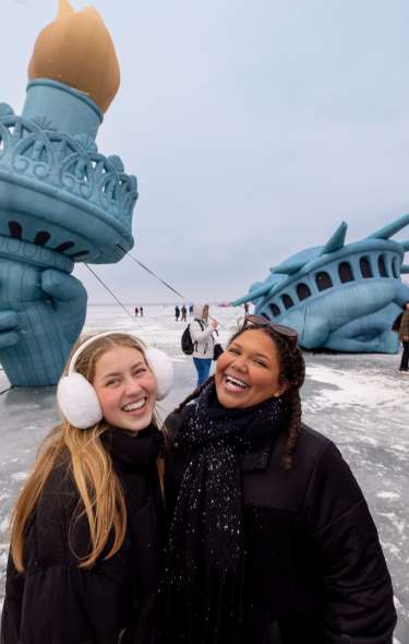Two young women are bundled up and laughing on the frozen lake in front of the large Statue of Liberty at the Winter Carnival.