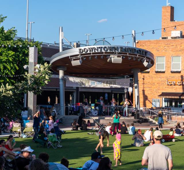 Looking at a distance towards a band playing on the stage at Main Street Square while attendees sit in lawn chairs scattered throughout the grass.