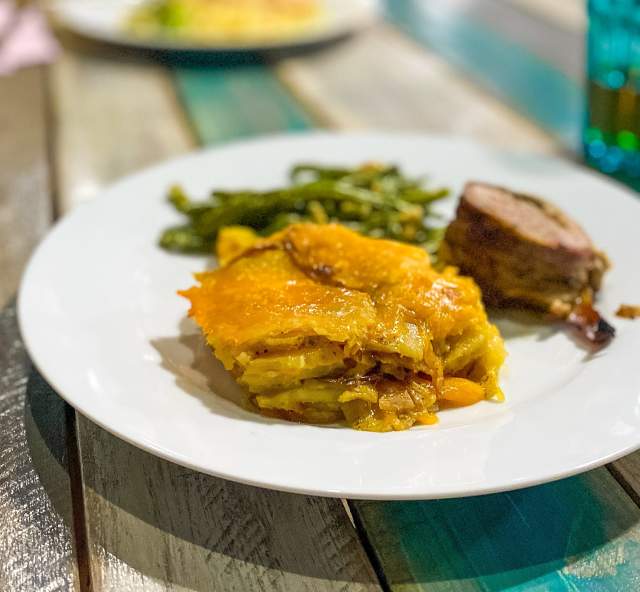 A plate of food on a rustic wooden table features golden potato gratin, green beans, and a stuffed meat roll. The setting is casual and inviting.