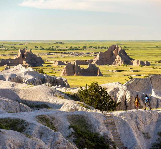 Two hikers walk along a rugged Badlands National Park trail surrounded by pale rock formations, grasslands, and distant buttes.