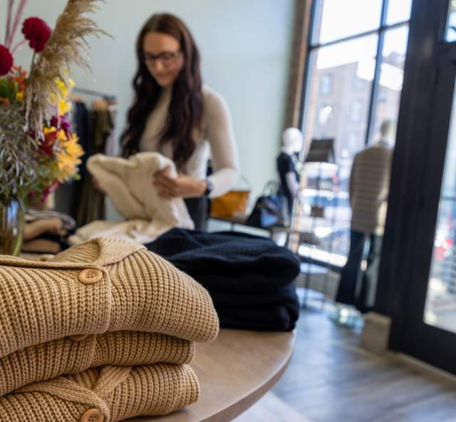 A young woman, slightly out of focus in the background, picks up a sweater in a well-lit boutique with huge windows. In the foreground are tan and navy blue cardigans on a table with a colorful flower arrangement in the middle of the table.