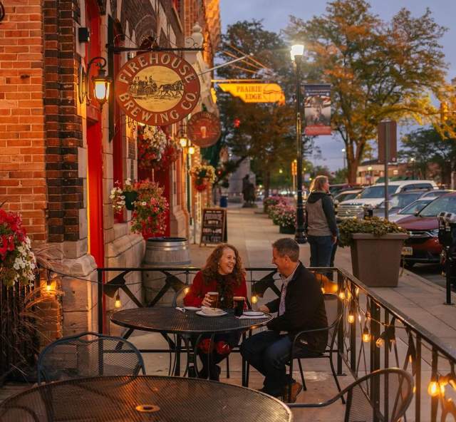A couple enjoys an evening on an outdoor patio at Firehouse Brewing Co. with brick walls and warm lighting, surrounded by flowerpots and streetlamps on a bustling city street.