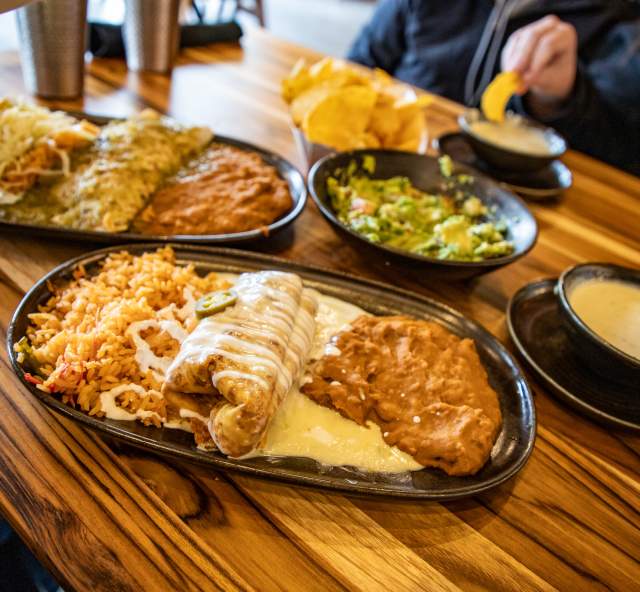 A queso-smothered burrito with Mexican rice and refried beans sits on a table at Ma Cualli Downtown, surrounded by tacos, guacamole, chips and dipping sauces.