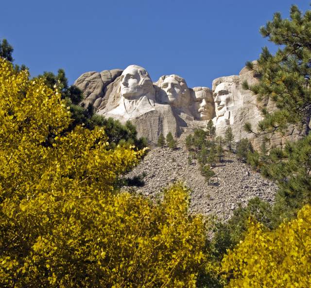 Mount Rushmore with four presidential faces carved into the granite, backed by a clear blue sky. Vibrant yellow autumn foliage in the foreground.