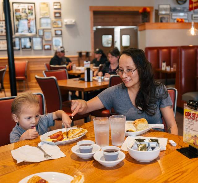 A mother and young child enjoy breakfast in a cozy diner. She helps him with a fork, surrounded by warm decor and other diners. The mood is cheerful.