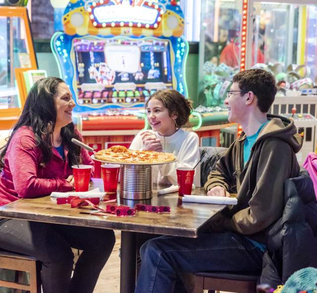 Family of three sitting around a table eating pizza with arcade games in the background at Press Start in Rapid City, SD