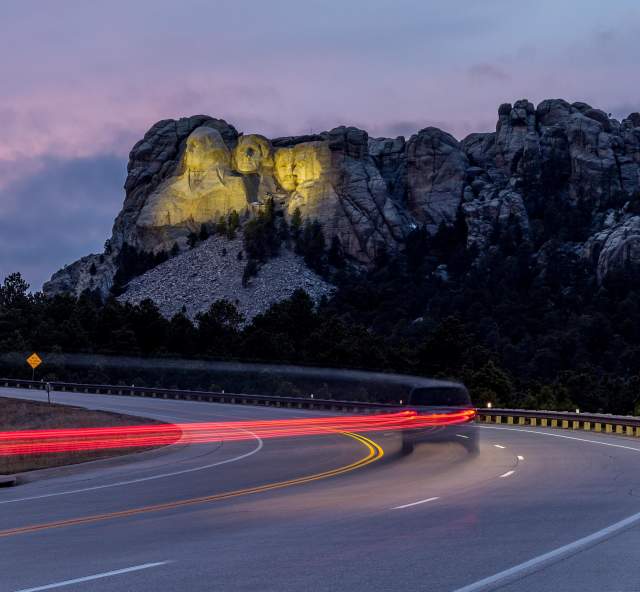 Mount Rushmore, lit up by yellow floodlights at dusk, sits in the background as a vehicle traveling down the highway is trailed by a long line of red taillights, created by a long-exposure photo.