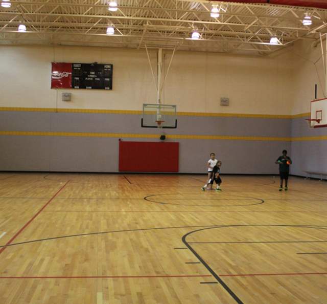 Indoor basketball gym with hardwood floors, multiple hoops, and overhead lighting, where three children stand near center court during practice.
