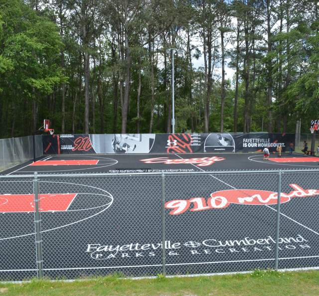 Fenced outdoor basketball courts at Smith Recreation Center with painted courts and Fayetteville-Cumberland Parks and Recreation branding, surrounded by trees