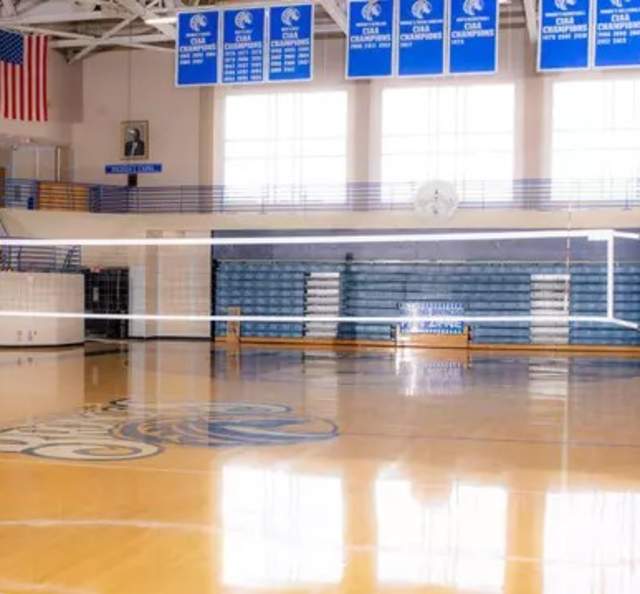 Indoor volleyball court at Fayetteville State University with polished hardwood floors, blue padded posts, and bleachers in the background beneath championship banners.