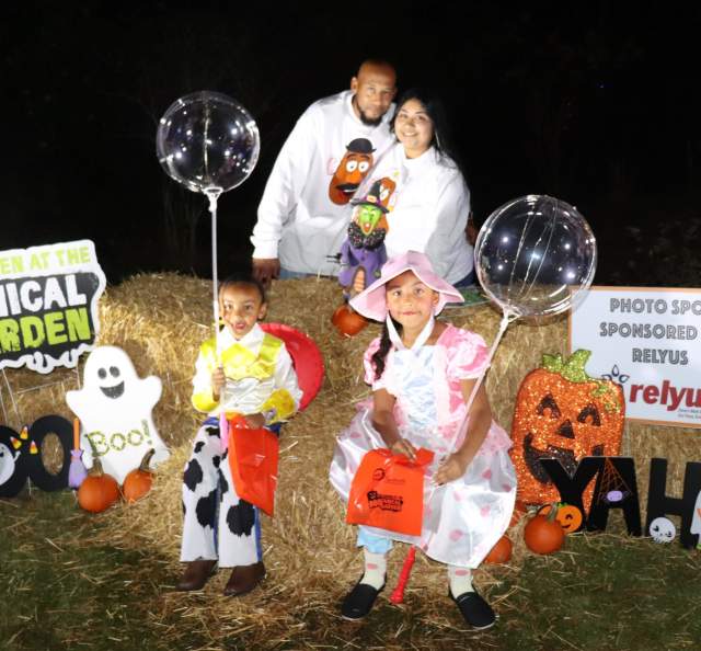 A family poses at a decorated Halloween photo spot during the Boo-tanical Garden event in Fayetteville. Two children in Toy Story costumes sit on hay bales holding clear light-up balloons and orange trick-or-treat bags, while two adults stand behind them smiling. The backdrop features pumpkins, ghosts, and a sign reading “Halloween at the Boo-tanical Garden.”