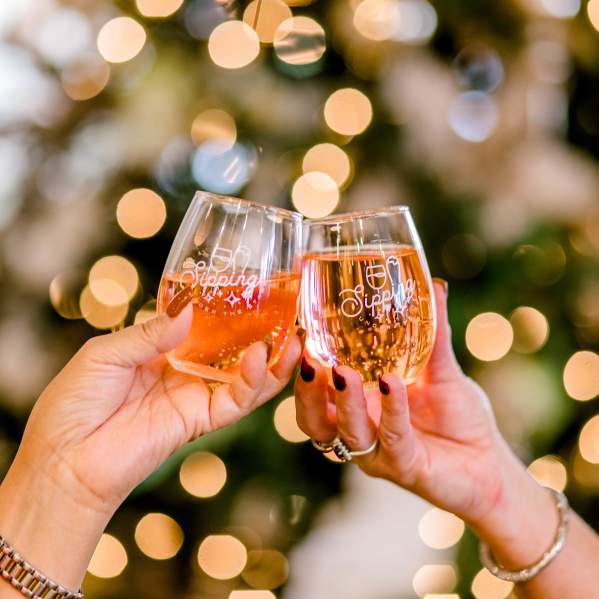 Two women tapping two wine glasses together for a cheers to the holidays in front of a lighted christmas tree in the background.