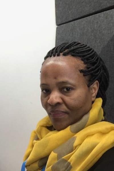 Close-up of a person with braided hair sitting indoors near acoustic wall panels, wearing a yellow hoodie, with part of a chair and a white wall visible in the background.