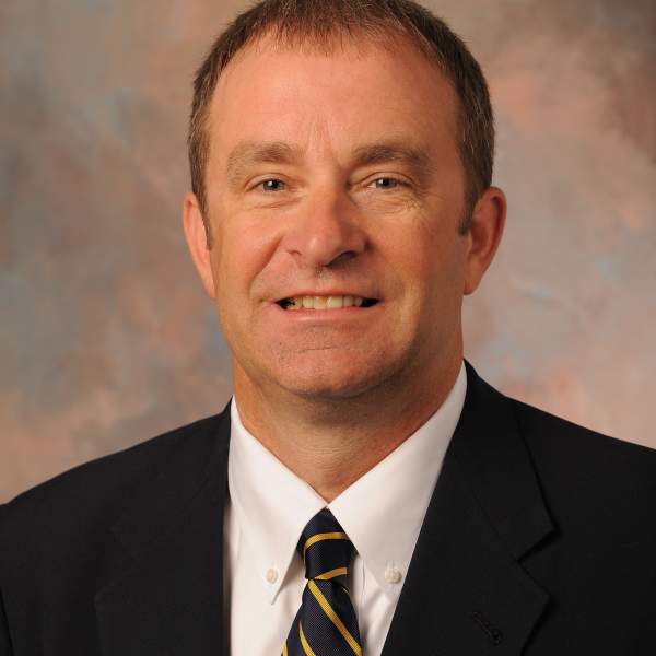 Professional headshot of a man in a dark suit, white shirt, and navy striped tie, smiling in front of a soft, multicolored studio backdrop