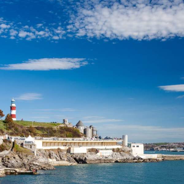 A coastal scene with a red and white lighthouse on a grassy hill, blue sky with scattered clouds, rocky shore, and calm blue sea creating a serene mood.