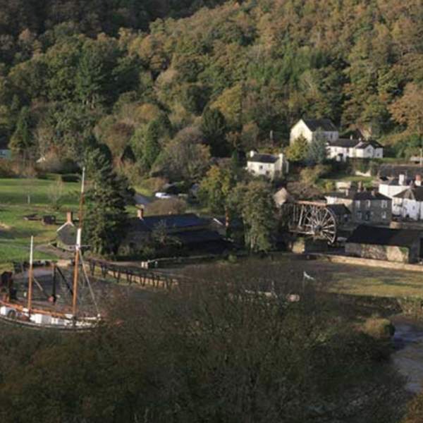 A scenic village nestled in a lush, forested valley. Features quaint cottages, a waterwheel, open green fields, and a docked ship, evoking tranquillity.