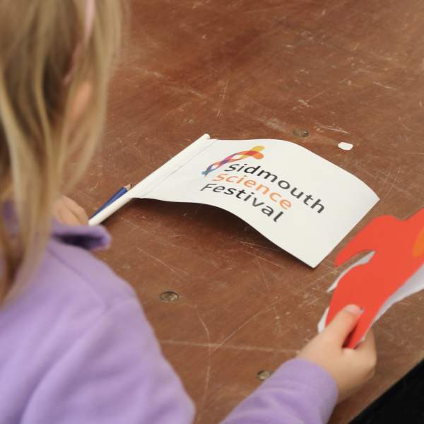 A child in a purple sweater holds a colorful paper rocket and a flag with "Sidmouth Science Festival" on a wooden table, conveying a playful, educational atmosphere.