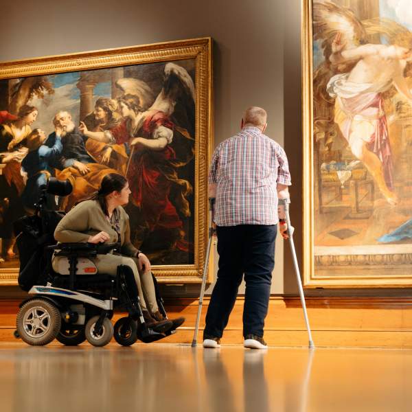 A woman in a wheelchair and a man using crutches viewing two large paintings inside Ferens Art Gallery. The spacious gallery features well-lit artwork and an accessible environment.