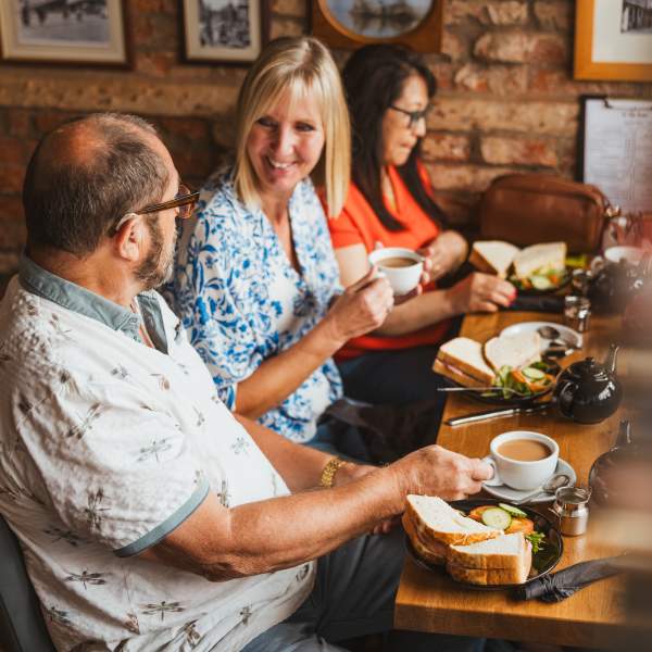 3 People sat a a table enjoying coffee and sanwichiches.