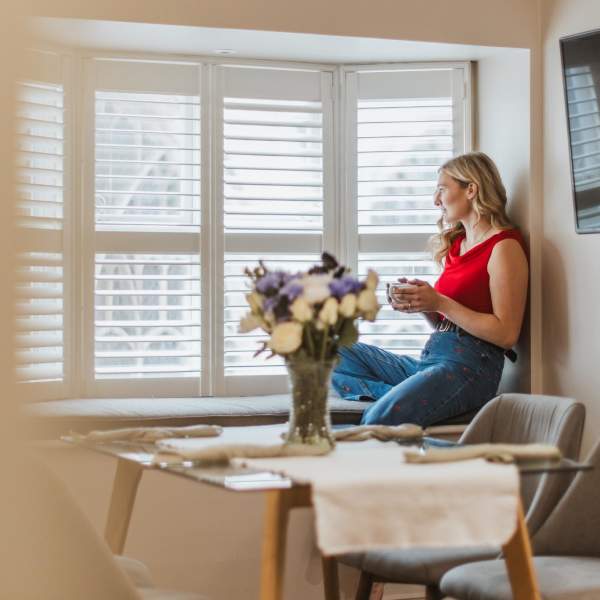 A girl in a red top sits on the inside of a set of bay windows, holding a mug and looking outside smiling. She has a table of flowers in front of her.