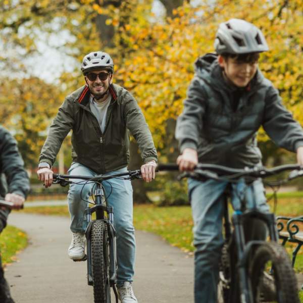 Three cyclists riding along a tree‑lined path in Hull’s Pearson Park during autumn, with fallen leaves and park benches visible