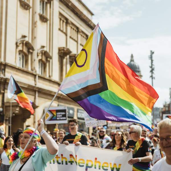Pride In Hull's parade walking down Carr Lane. The sun is shining, the crowd is smiling and dressed in shorts and t-shirts. A group of people holding a rainbow Smith+Nephew banner are in the foreground with a brightly dressed woman waving an Intersex Inclusive pride flag. Many other pride flags can be seen in the background.