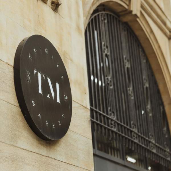 The sign for Trinity Market. The black arched entrance is to the right. The sign is black with a white "TM" in the centre in a sleek font. "Trinty Market" curves around the cirular edge of the sign.