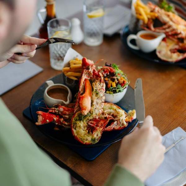 Man sat at a table tucking in to a delicious Bridlington Bay lobster. The men has his back to the camera and the lobster is in the centre of the image. It is filled with a herby sauce and is accompied by a salad and chips. Across from him someone else is eating the same thing.