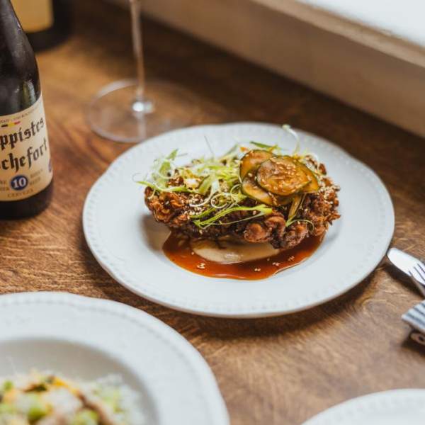 Small plates on a wooden table with a bottle of wine to the left. The plates are filled with well presented food and garnished with micro greens and a drizzle of red sauce.