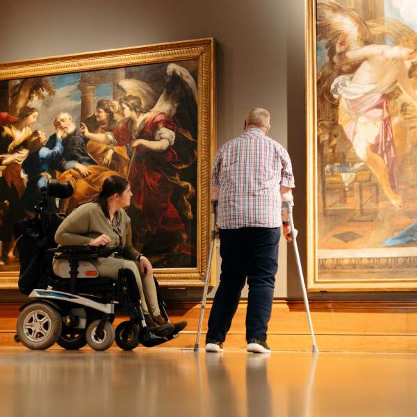 Two large and ornate paitings hang in Ferens Art Gallery. A couple are admiring the art. The woman has dark hair and is in a motorised wheelchair. The gentleman is using crutces and wears a plaid shirt.