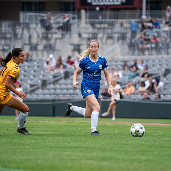 Two female soccer players, one in yellow and the other in blue, compete for the ball on a lush green field, with spectators in the background.