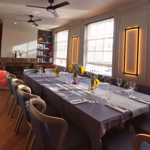 Dining table set up for private dining at Market House Cambridge. The table has a dark tablecloth with white place settings & posies of flowers in the centre of the table/ The room is light & bright with large windows to the right of the image