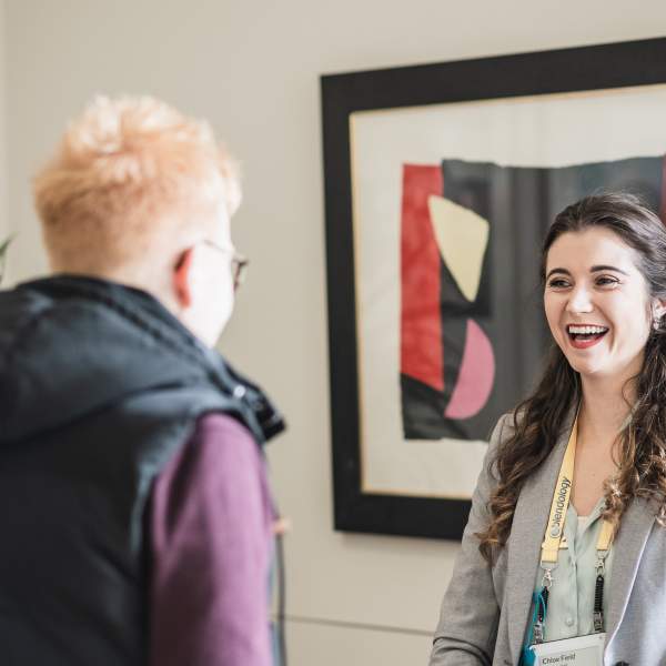 A young white woman with long wavy brown hair smiling in front of an abstract art picture whilst chatting to another white woman who has her back to the camera