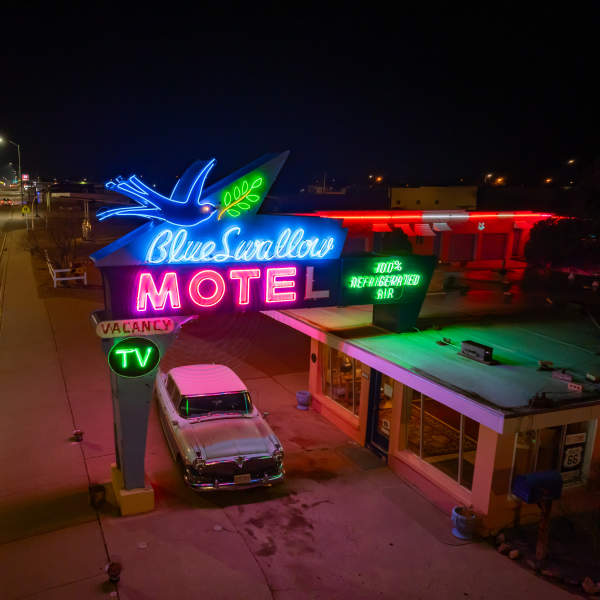 a classic 50's car sits outside of a motel beneath a warm neon sign, the sign reads "Blue Swallow Motel, 100% refrigerated air, TV"